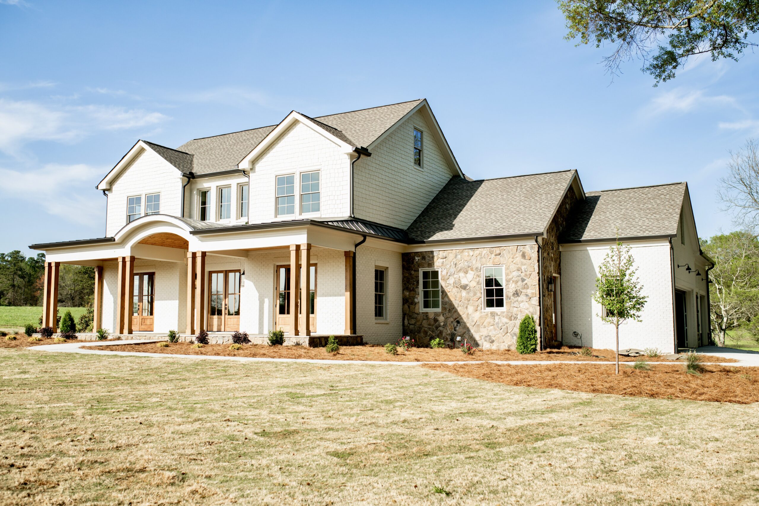 Modern white farmhouse exterior with covered front porch, stone accent wall, dark shingle roof, and attached garage on a landscaped lot