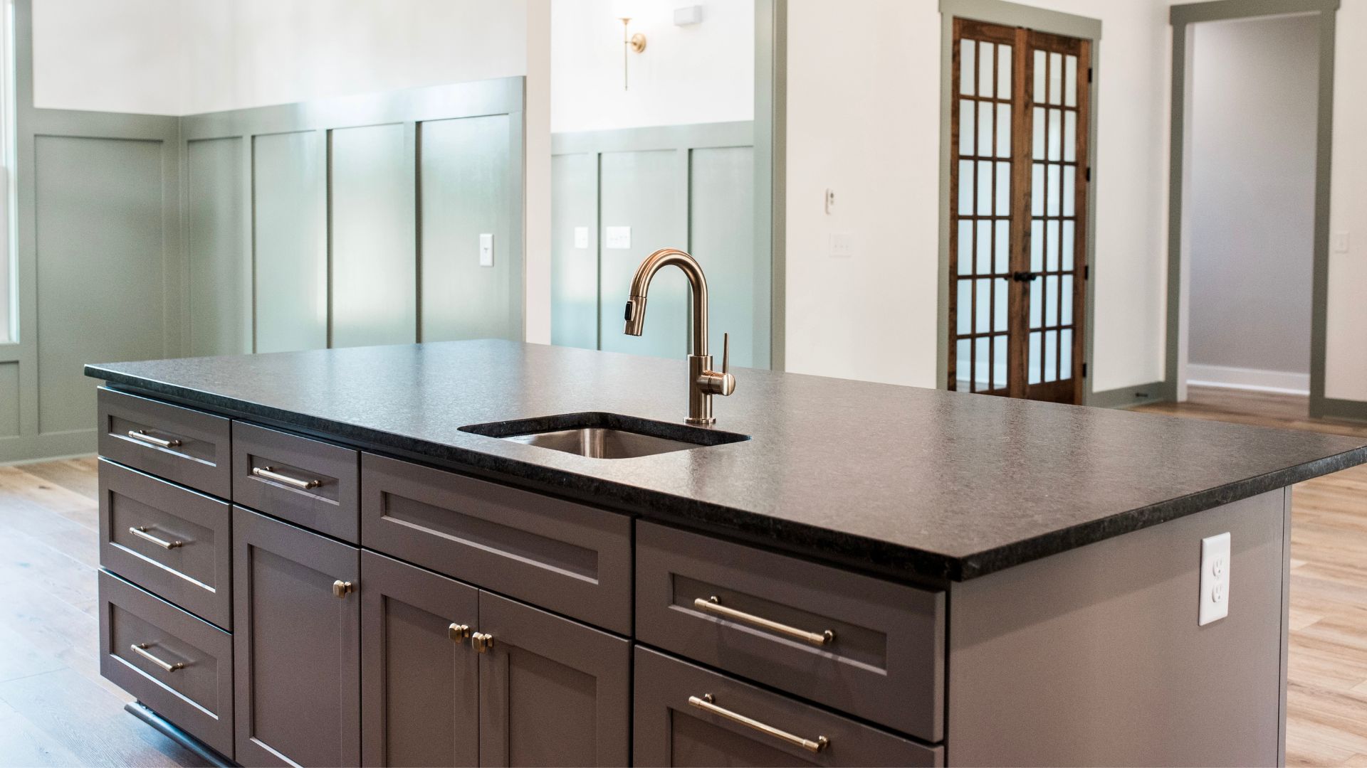 Large kitchen island with dark cabinetry, black granite countertop, undermount sink, gooseneck faucet, and wood French doors in background