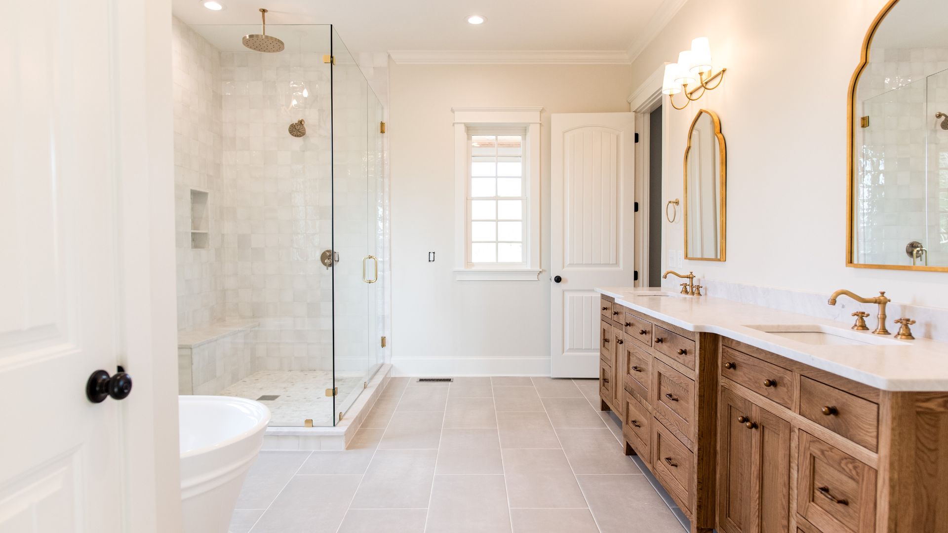 Bright master bathroom with glass walk-in shower, double vanity with wood cabinets, white countertops, brass faucets, and arched mirrors
