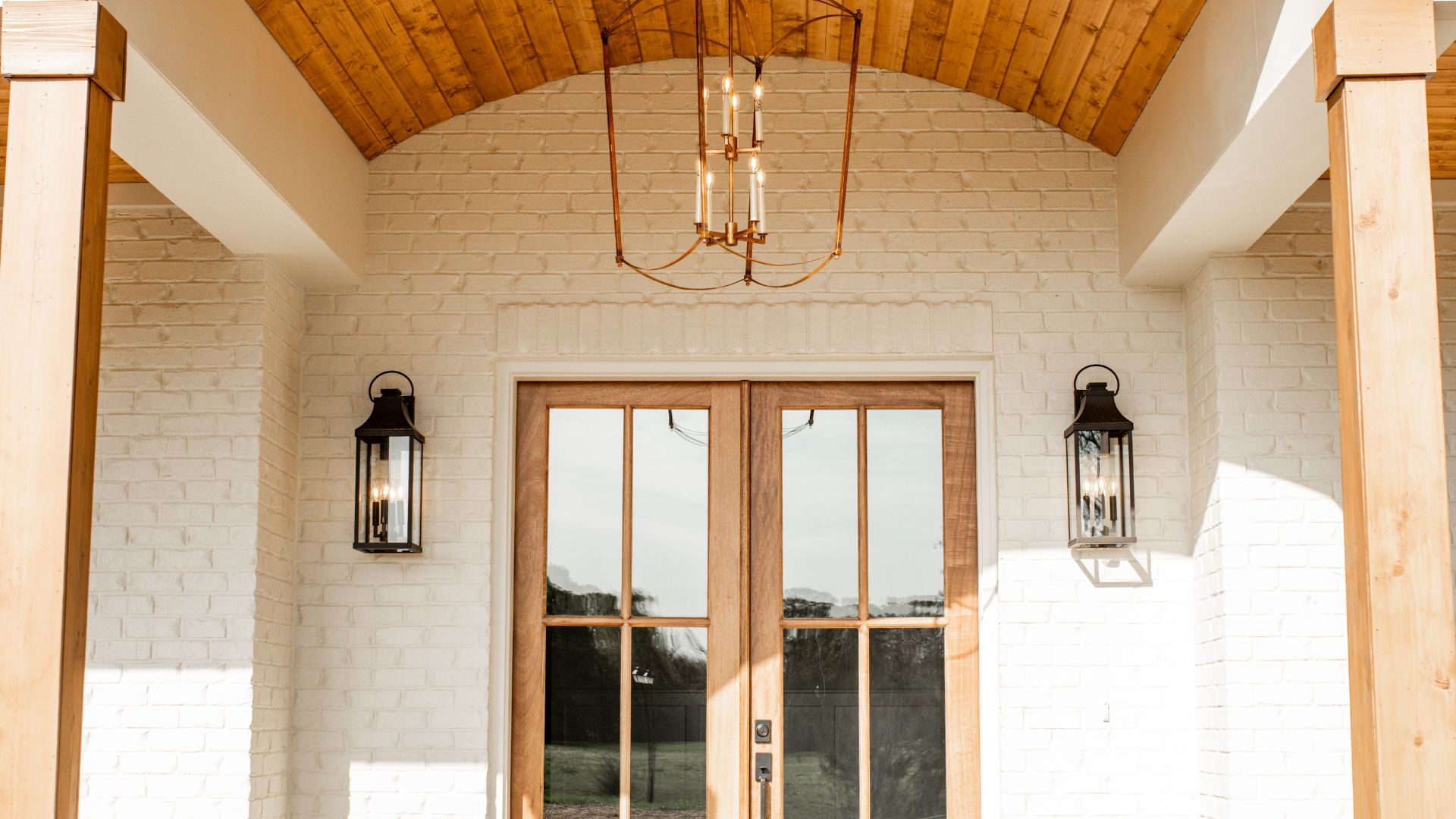 Front porch entry with arched wood ceiling, white brick walls, glass double doors, black lantern sconces, and hanging chandelier