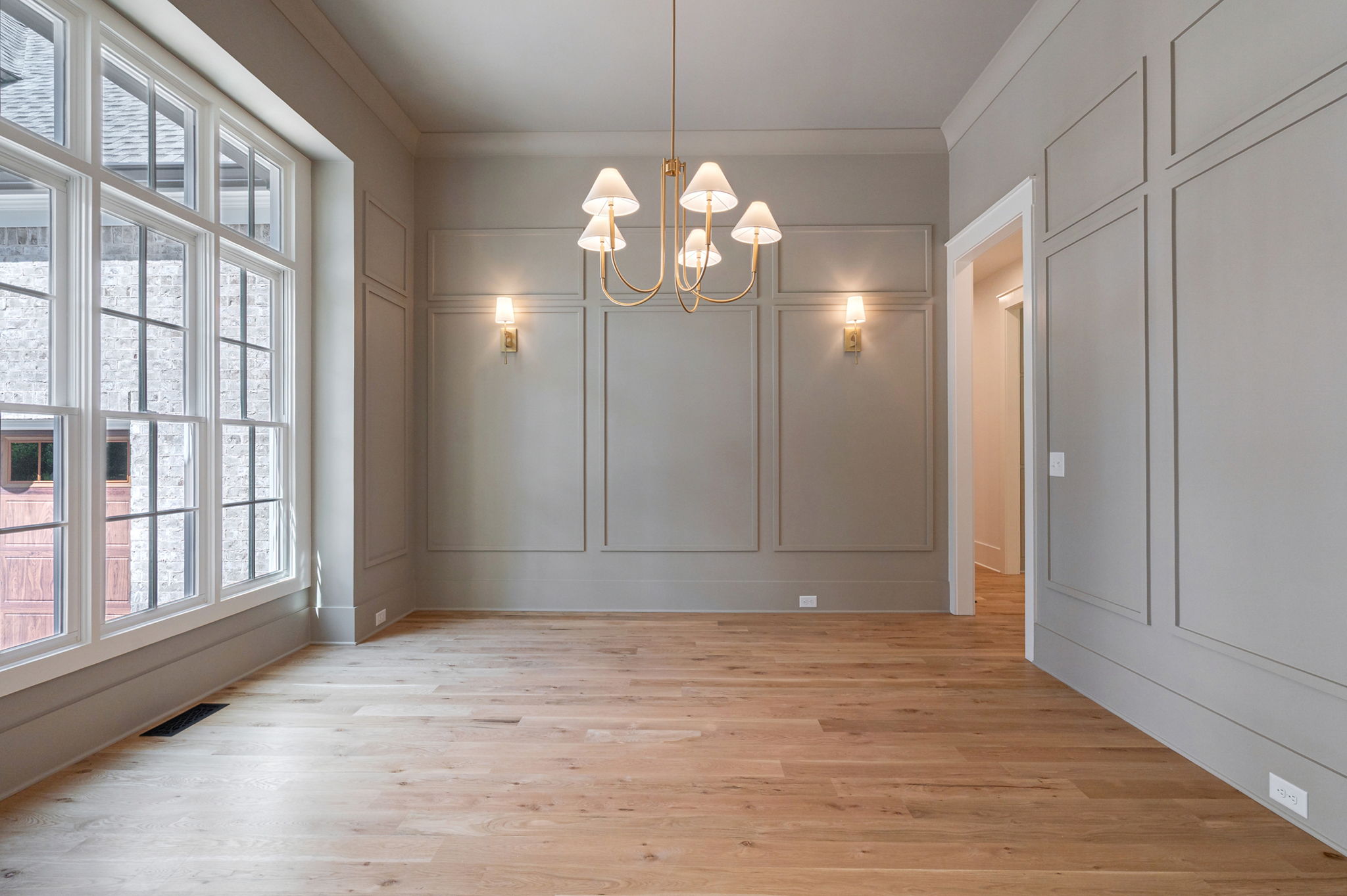 Empty dining room with light gray wall paneling, brass chandelier and sconces, wide hardwood floors, and large grid windows
