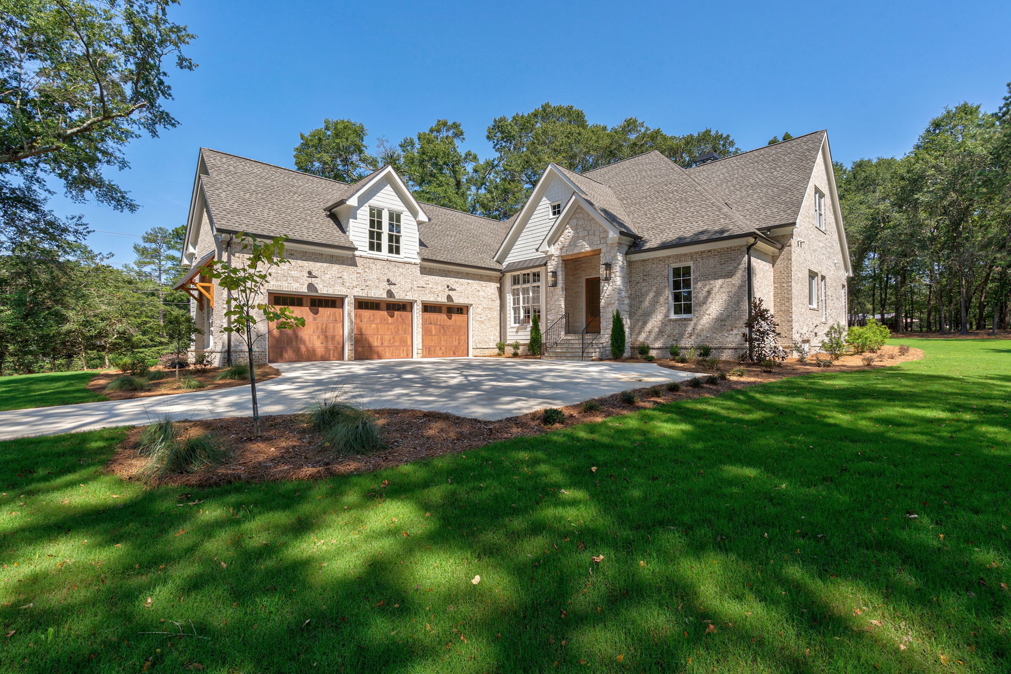Custom brick home exterior with three-car garage, stone entry, concrete driveway, and landscaped front yard under a clear blue sky