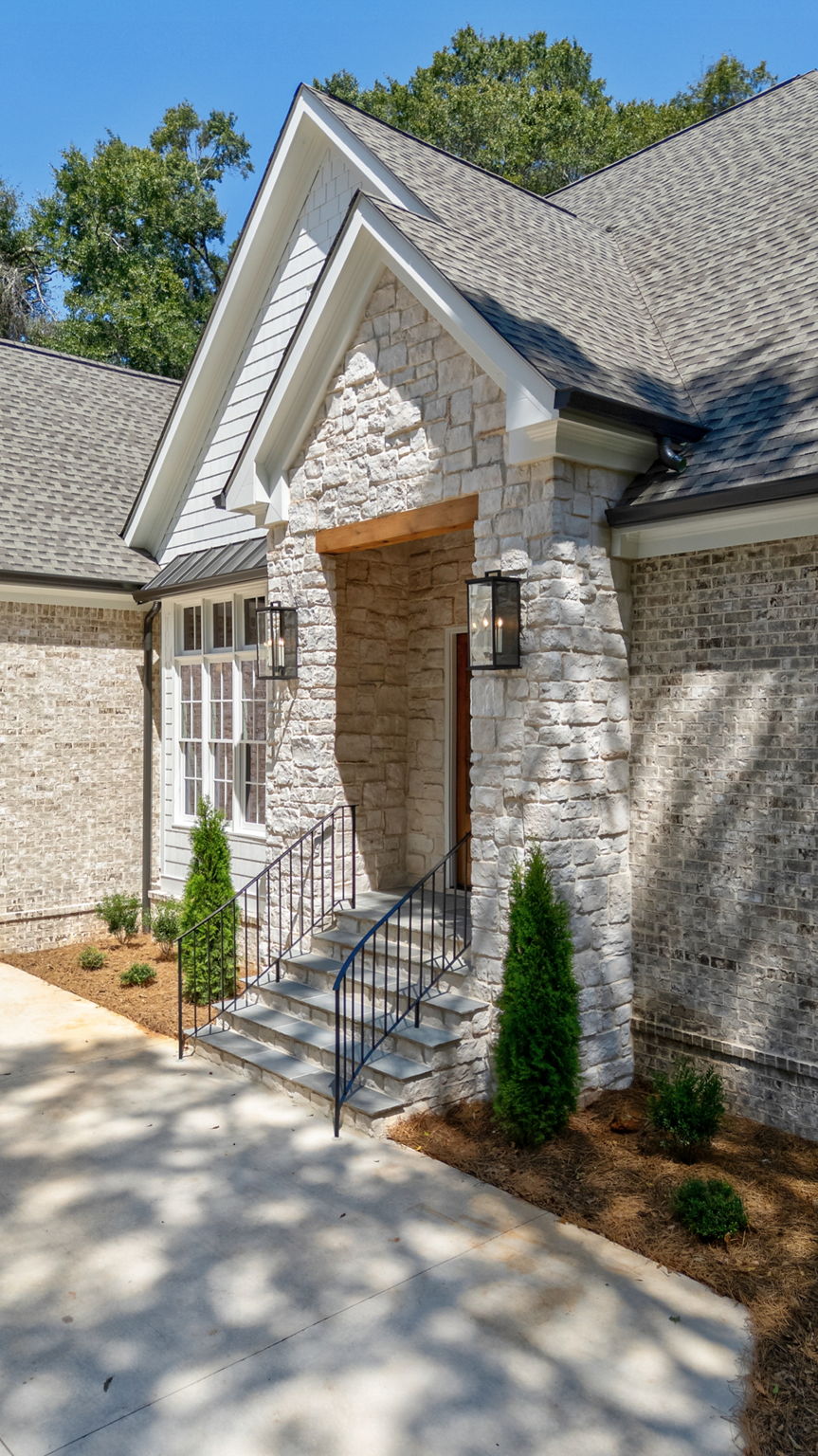 Stone entryway with covered front porch, gabled roofline, black lantern lights, concrete steps with iron railings, and fresh landscaping