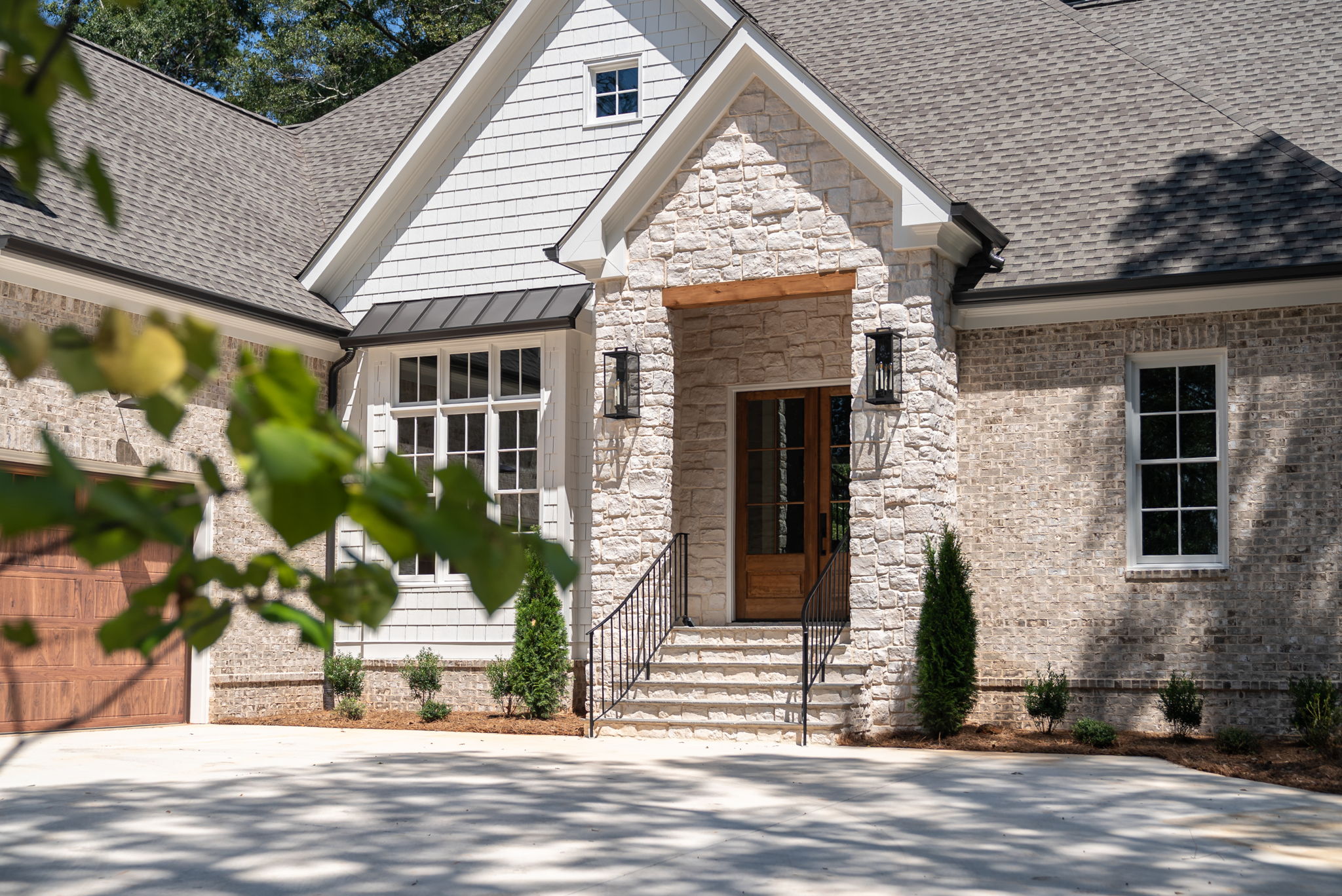 Front entry of a custom brick home with a stone gable porch, wood front door, black lantern lights, and steps with iron railings