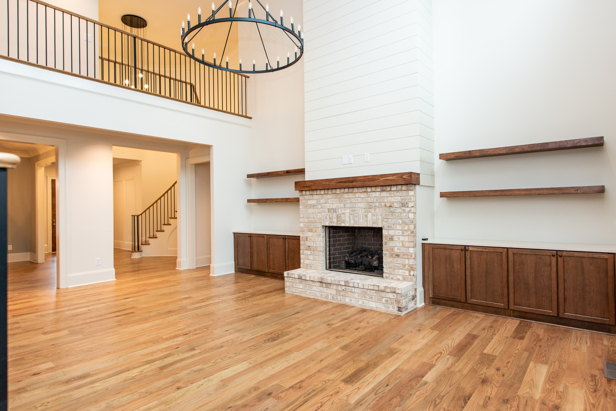 Bright open-concept living room with white shiplap fireplace, brick hearth, floating wood shelves, built-in cabinets, hardwood floors, and oversized round chandelier